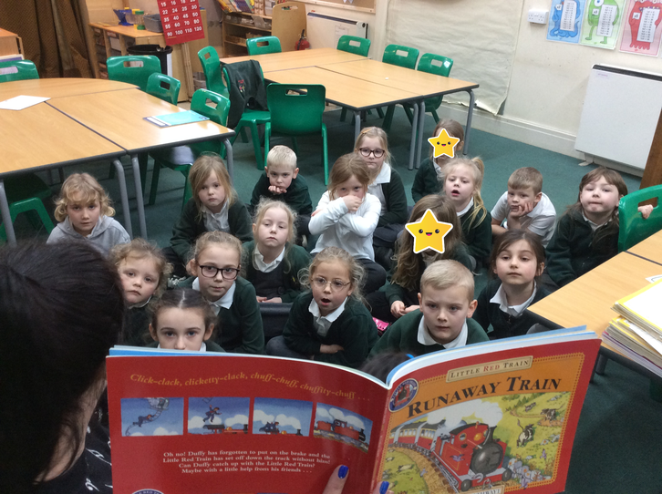 Primary school children in green uniforms sit on the classroom floor, attentively listening to a 'Runaway Train' storybook reading. Some faces have star stickers.
